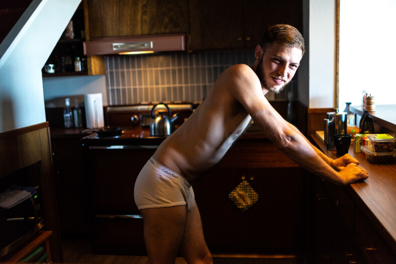 A man with a beard wearing nothing but Calvins leans on the counter in his kitchen, posing for the camera.
