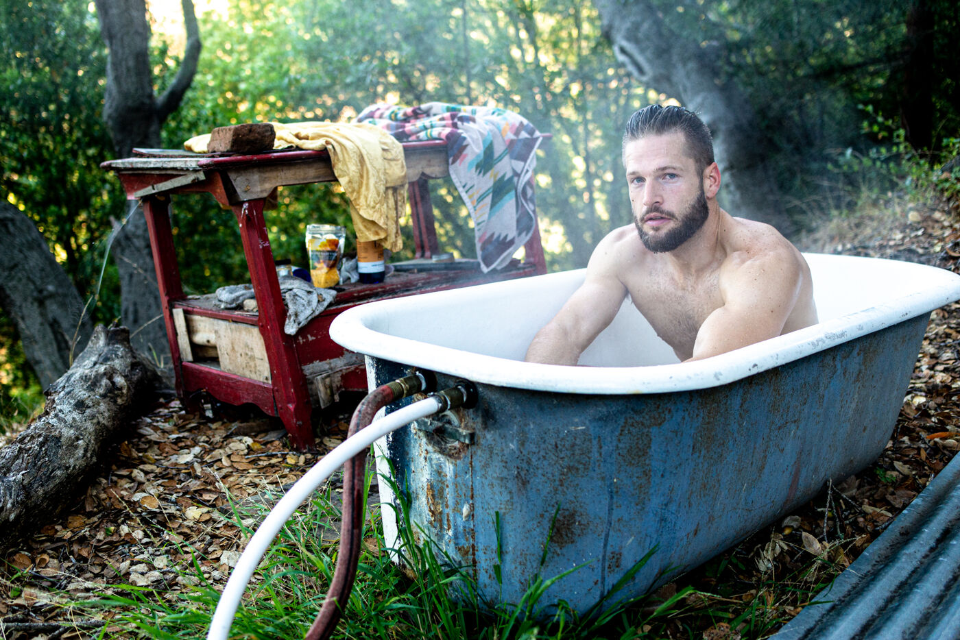 A shirtless man sits in a bath tub in the woods. 