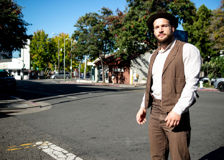 A bearded man in a striped vest, shirt and trousers, wearing a fedora poses for the camera on the street.