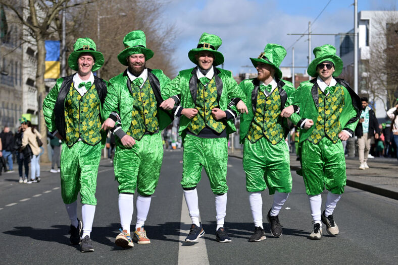 5 men in St. Patty's Day costumes walking down the street arm in arm.