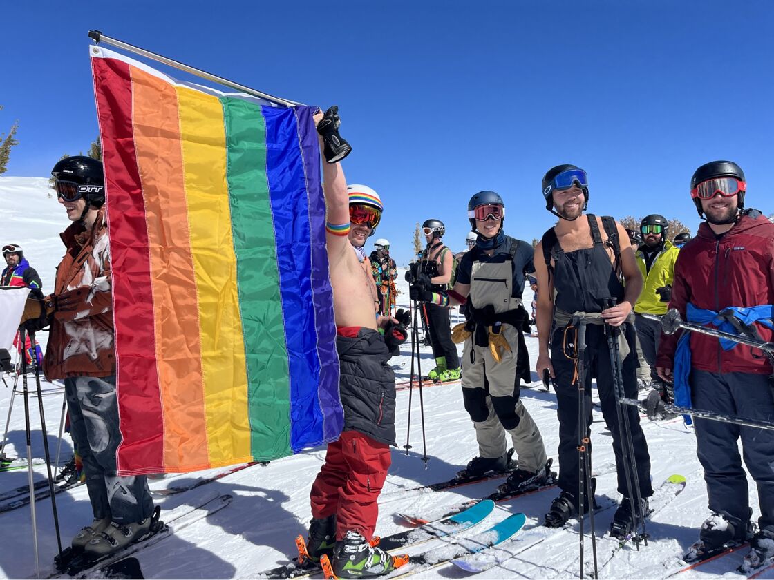 Skiers on the mountain getting ready for the rainbow run. One of them is holding a large rainbow pride flag