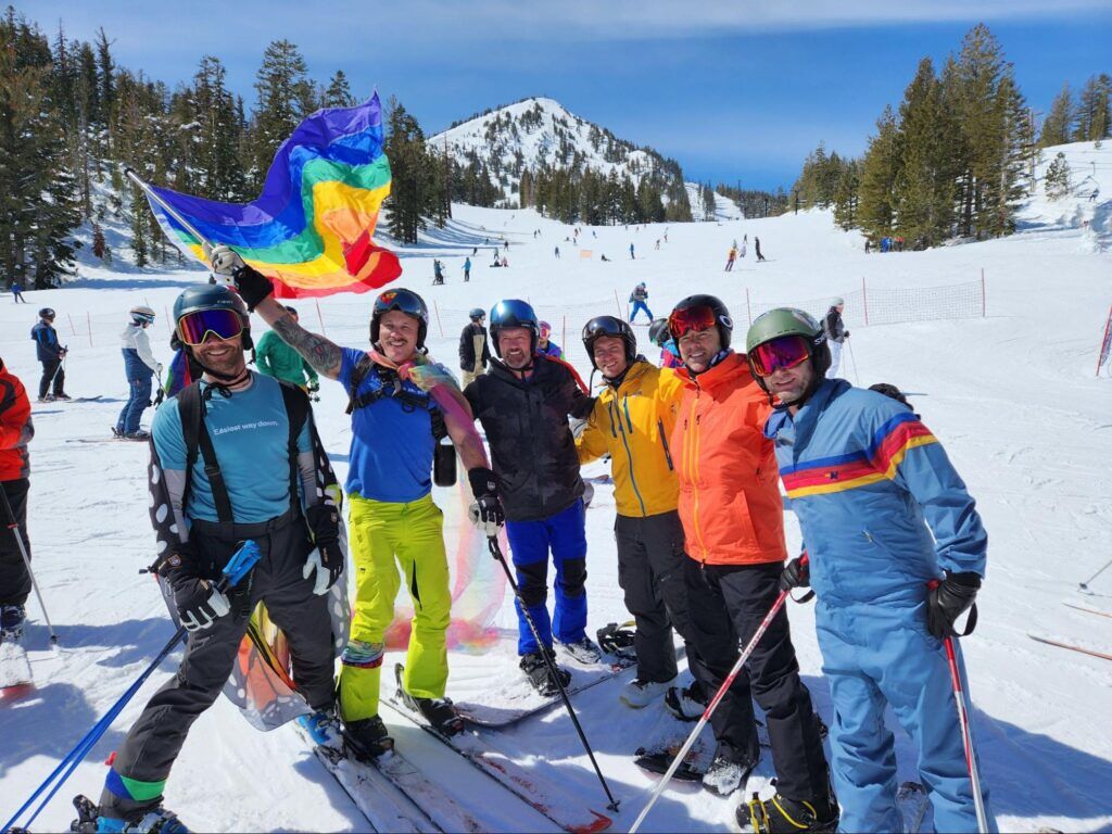 Skiers on the mountain getting ready for the rainbow run. One of them is holding a large rainbow pride flag