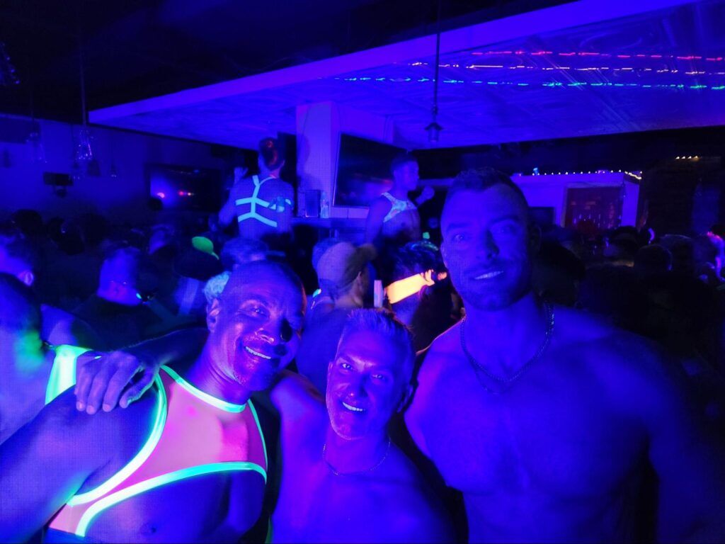 Three men posing for the camera in black light, one of them is wearing a glow in the dark chest harness. 