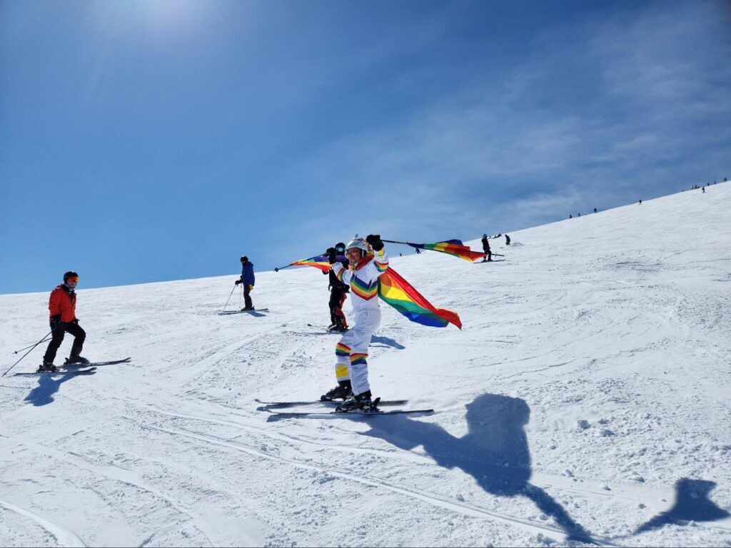 A skier wears a rainbow Pride flag as a cape as they go down the mountain.