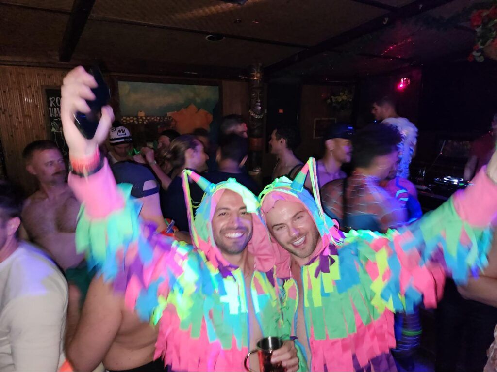 Two guys in colorful onesies dancing in the crowd at a bar.