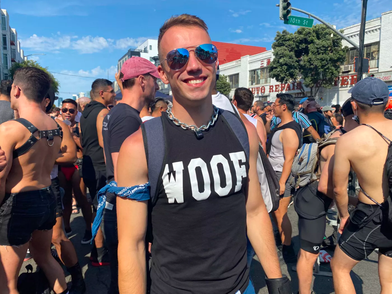 A guy wearing sunglasses, a chain collar, a blue bandana around his bicep, and a shirt that reads "woof".