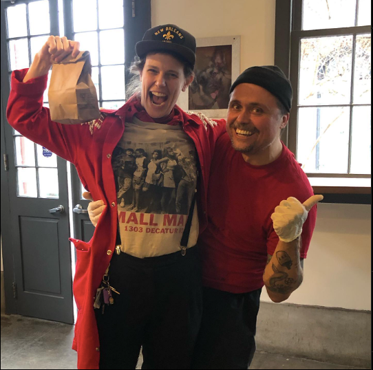 A patron dressed in red with a black beanie holds up their bagel while the worker also wearing red and a black beanie gives a thumbs up. 