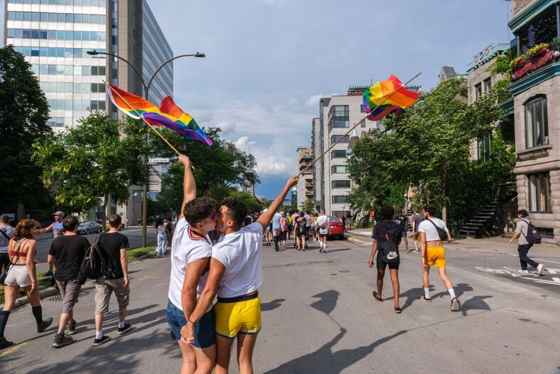 Two men standing in the street, holding rainbow pride flags, kissing at the Montreal Pride Parade