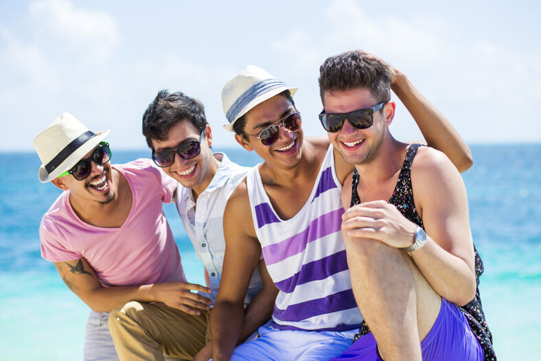 Four guys in summer clothes and sunglasses hanging out on the beach.