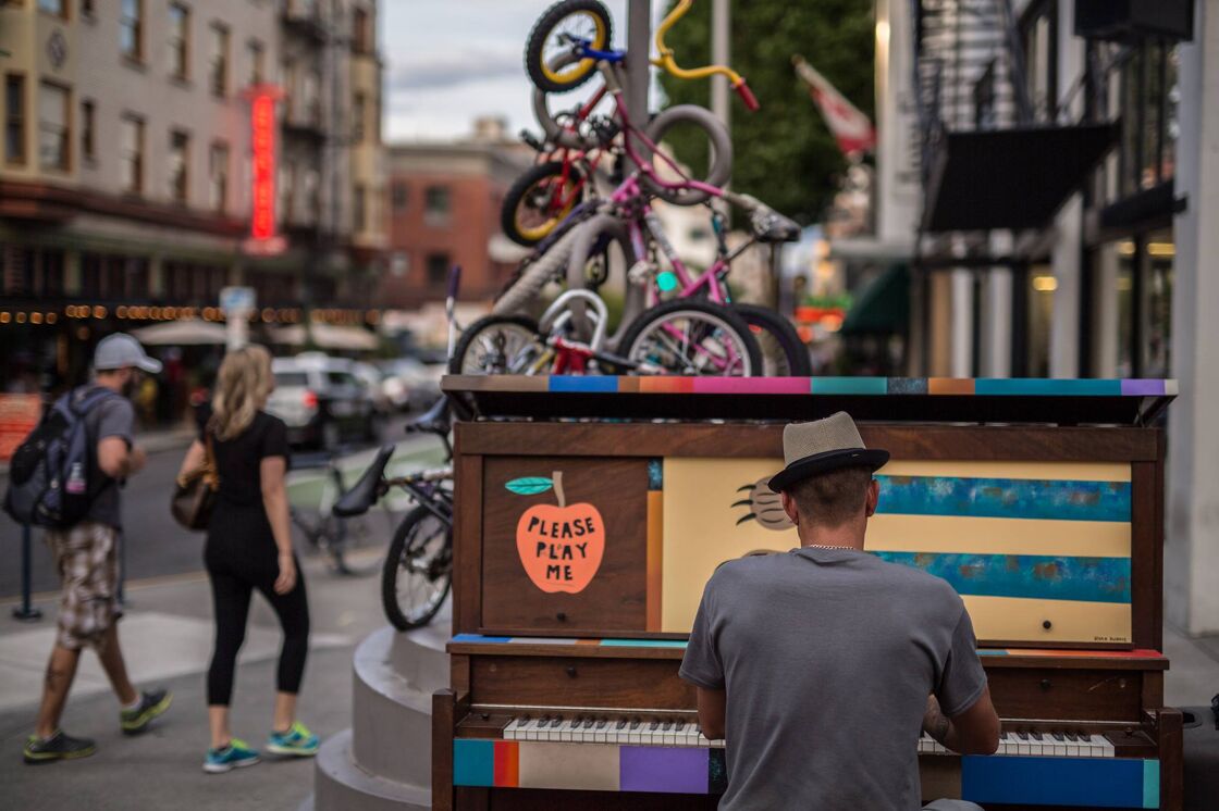 A man plays a piano on a street corner. The piano has a sign on it that says "Please play me."
