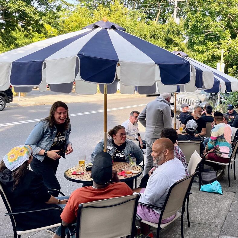 People gossip and drink outside under an umbrella at Sissy Bar Portland.