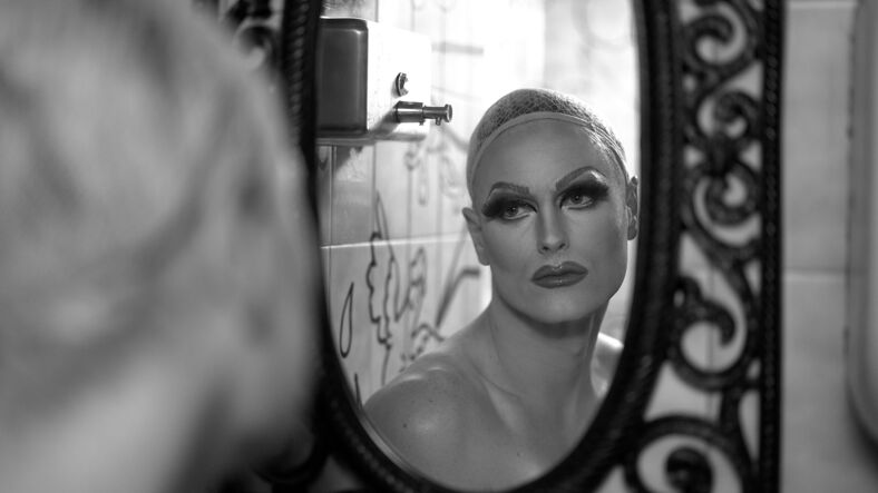 A draq queen looks into a bathroom mirror in full makeup.