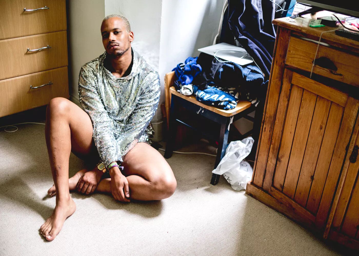 A man wearing a sequin hoodie sitting on the floor of his bedroom. 