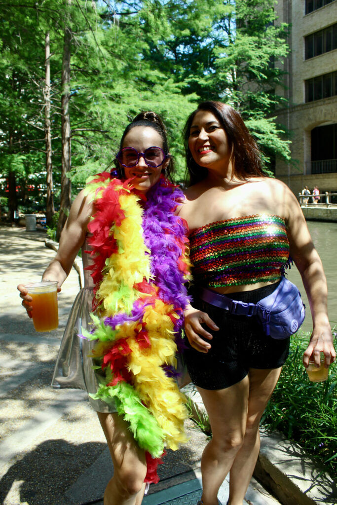 Two women adorned in rainbow attire stand in the shade. One wears a multi-colored feathered boa, the other wears a rainbow sequined tube top