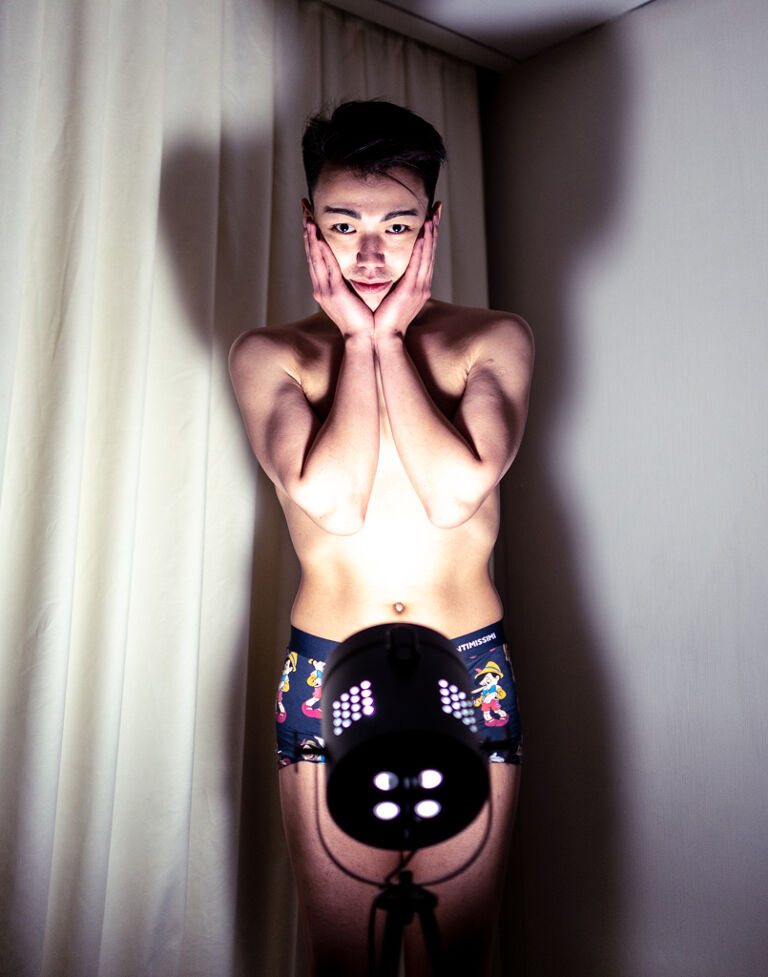 A shirtless man in boxers stands in front of a spotlight holding his face.