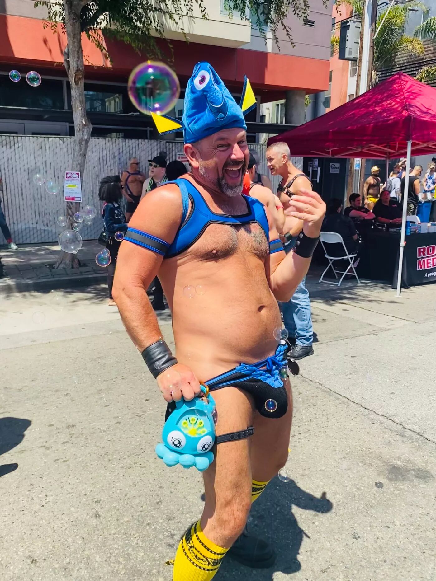 A nearly nude man sporting a Dory hat and blue jockstrap blows bubbles in the street