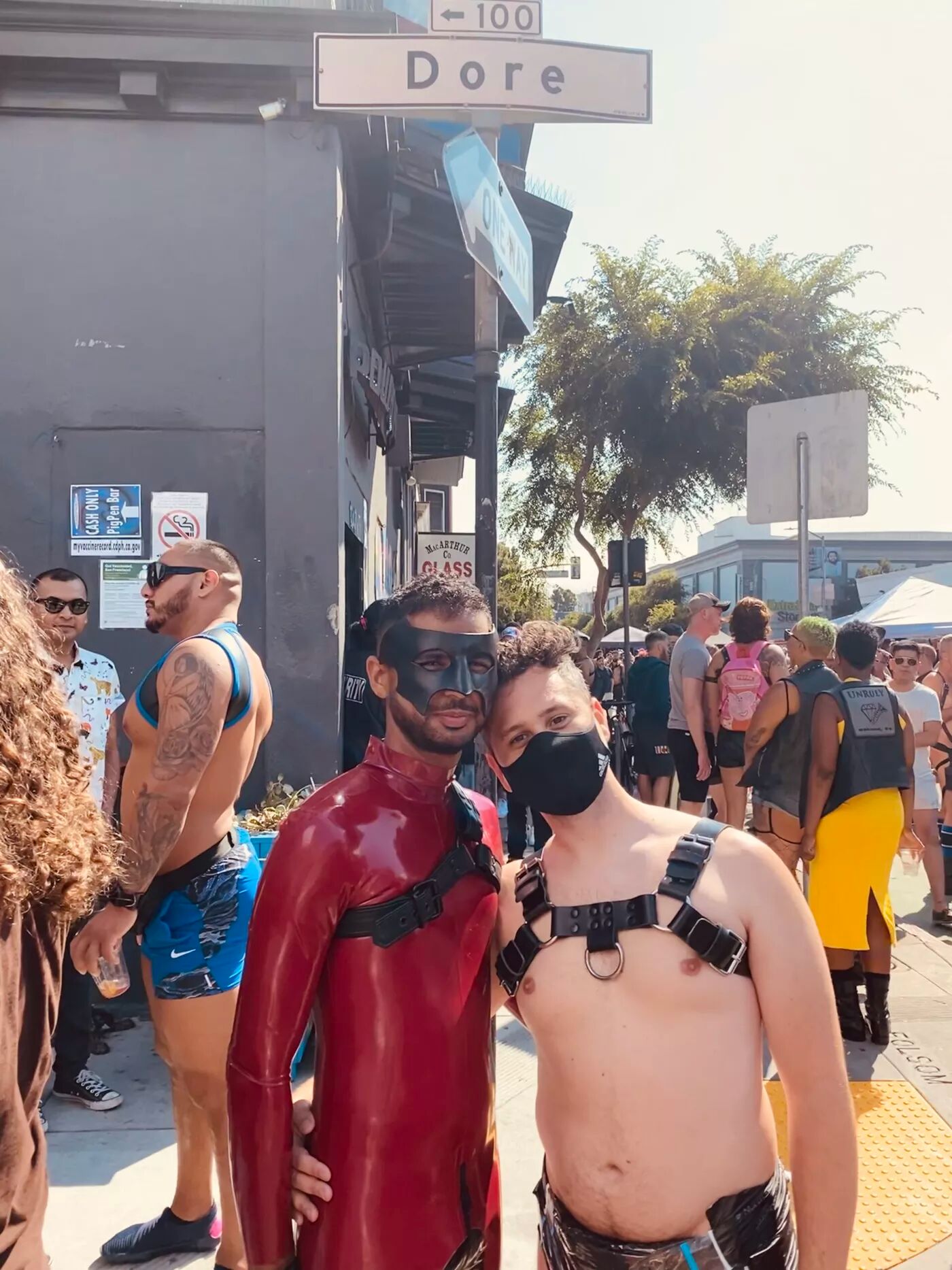 Two men stand in a busy street. One wearing a leather harness and face mast, the other dawning a full body red latex suit and batman mask