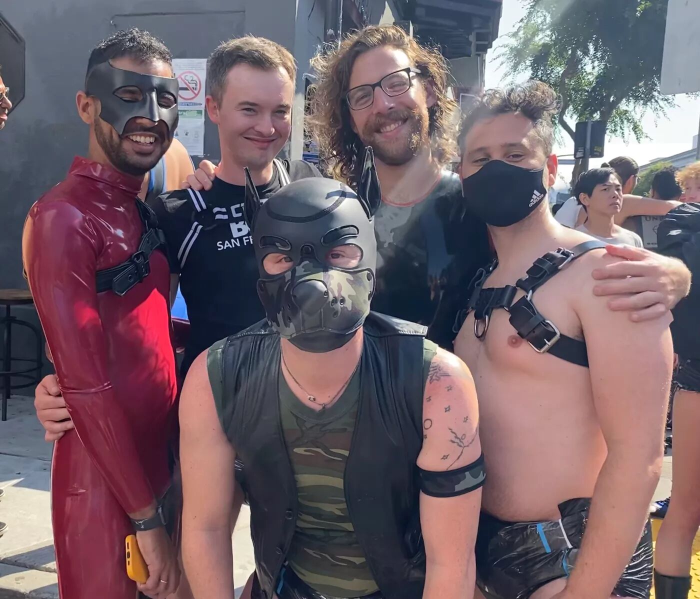 A group of men adorned in various kink and fetish attire stand together smiling at the camera