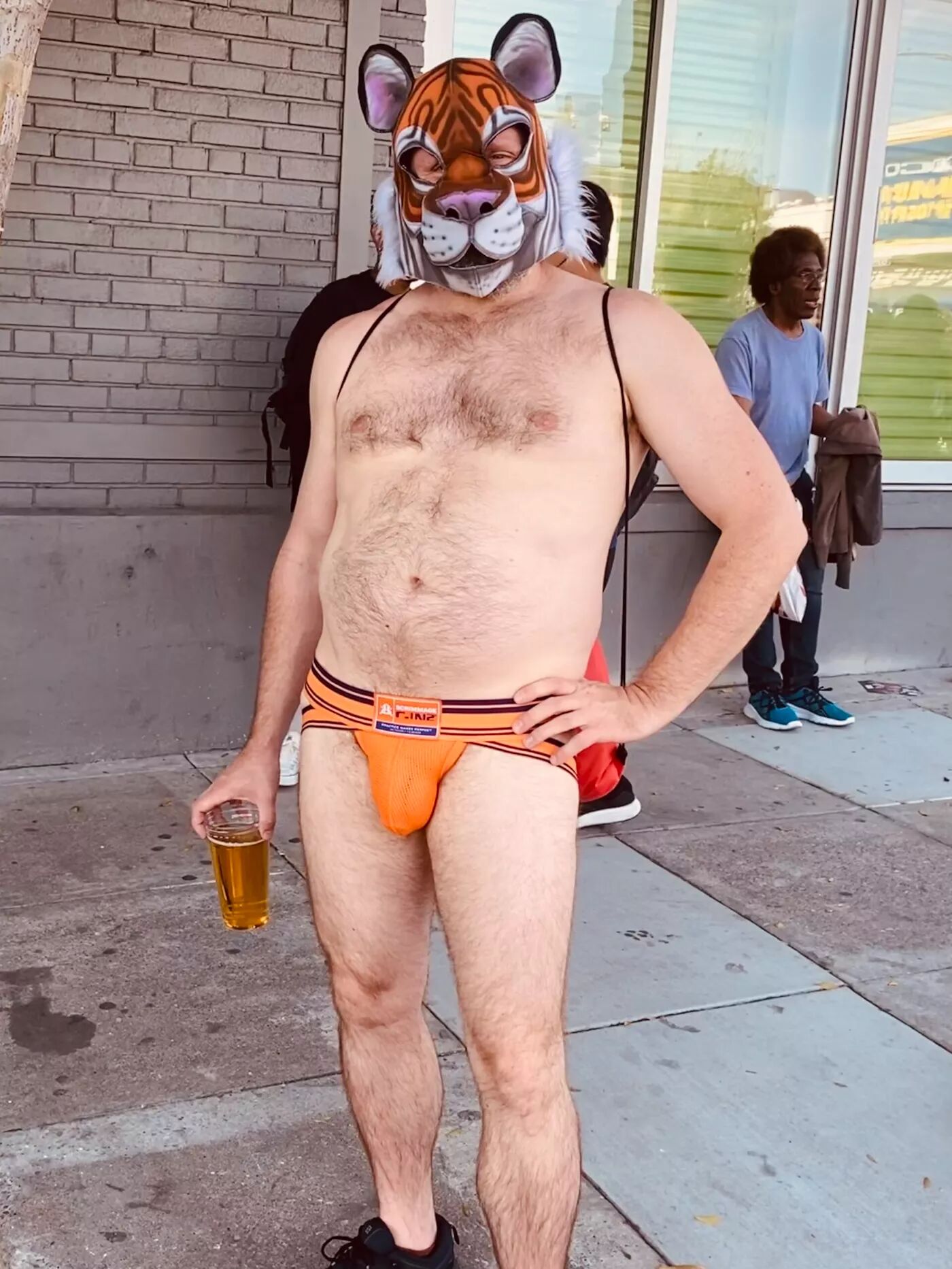 A man in an orange jock strap, wearing a tiger mask stands proudly while holding a full beer at his side