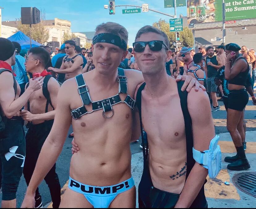 Two shirtless men stand in a busy street. One wears a leather harness and jockstrap, the other sunglasses and suspenders 