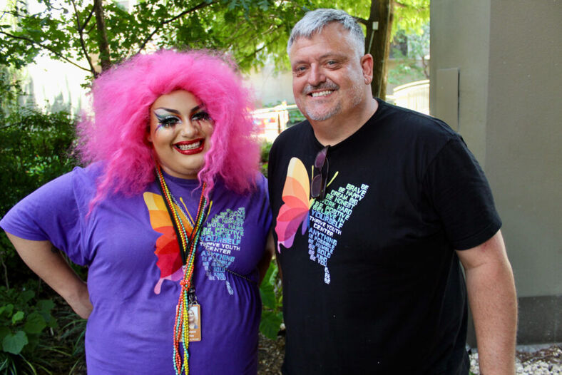 Two people stand smiling. One has wild pink hair and drag makeup