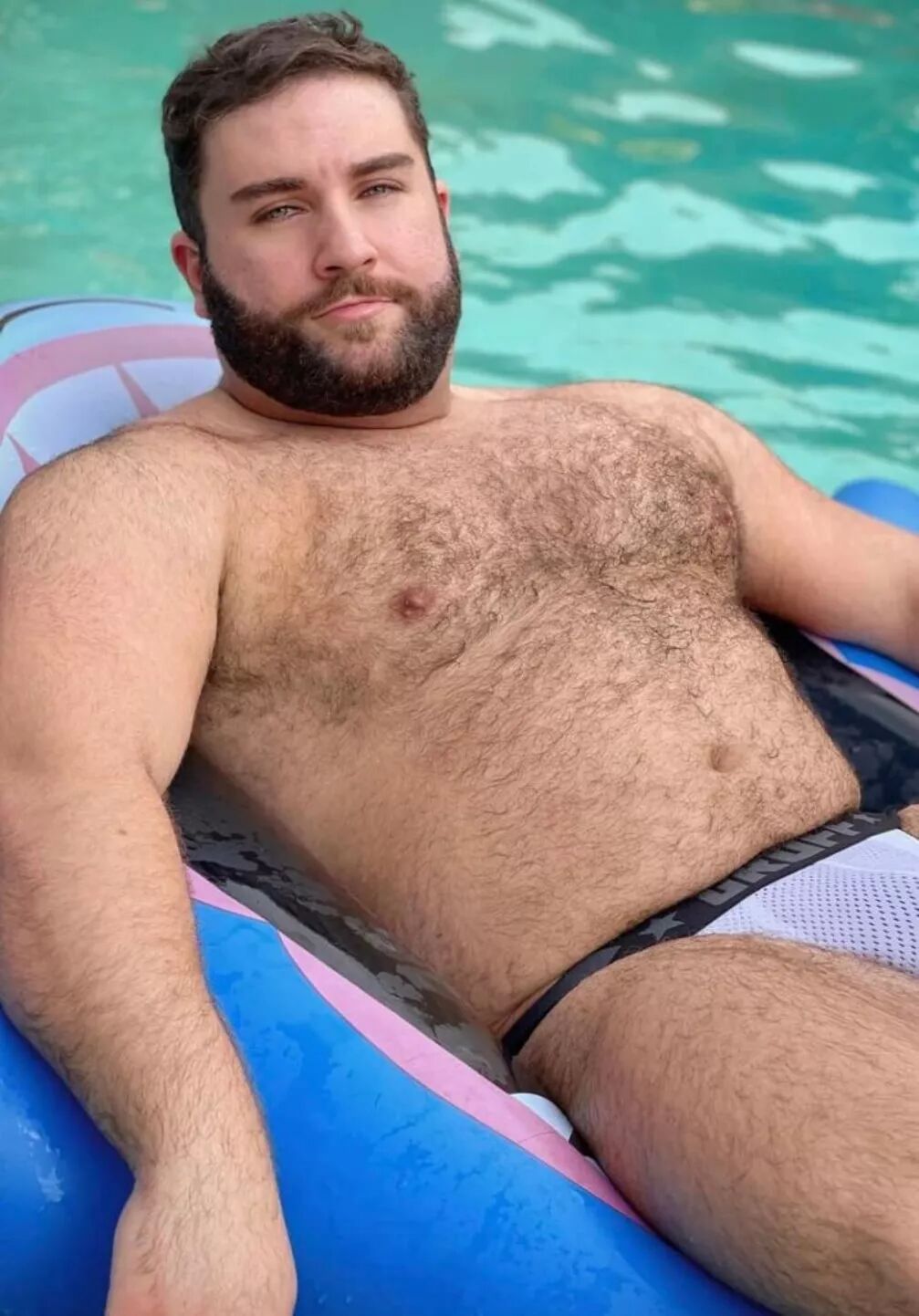 A hunky bear lounging in a pool floaty wears a white mesh jockstrap