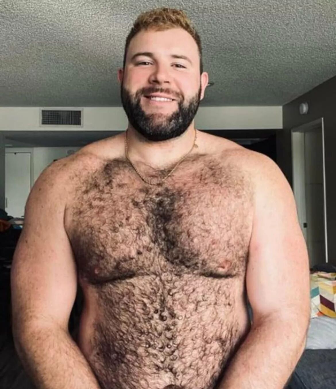 A smiling hairy bear smiles at the camera while sitting in an apartment