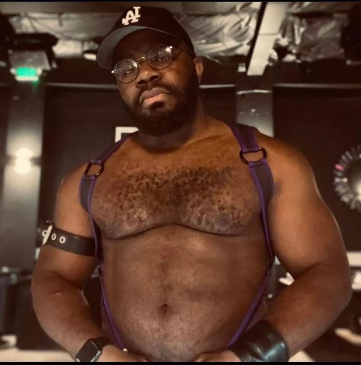 A hunky bear wearing a harness, baseball cap and round glasses