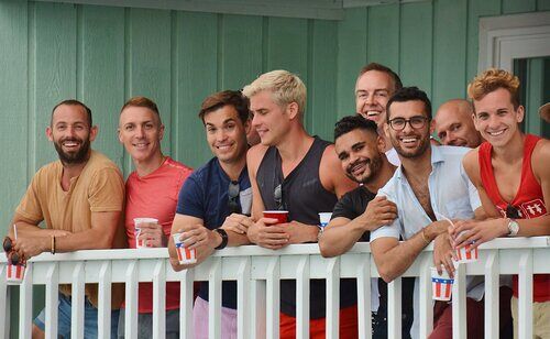 A group of smiling men lean over a wooden balcony railing