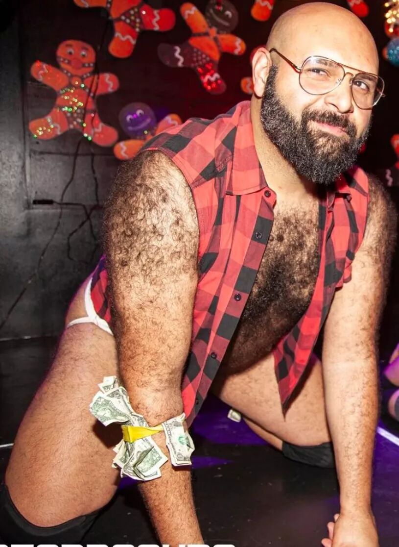 A bearded bear wearing a sleeveless lumberjack shirt and round rimmed glasses gets on all 4s