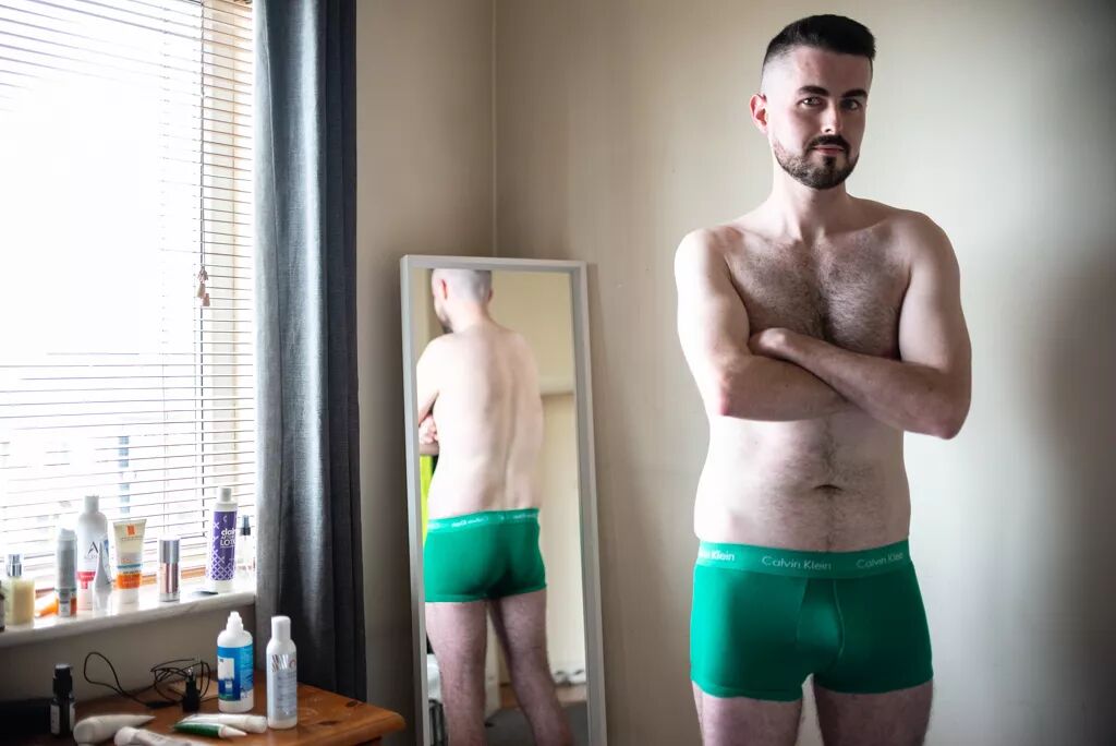 A man stands in his bedroom in front of a mirror, wearing bright green boxer briefs