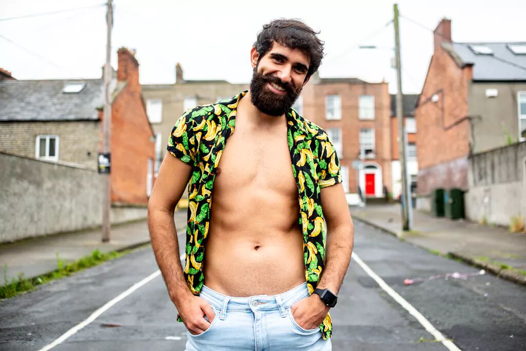 A smiling bearded man stands with his colorful shirt open in the middle of a street