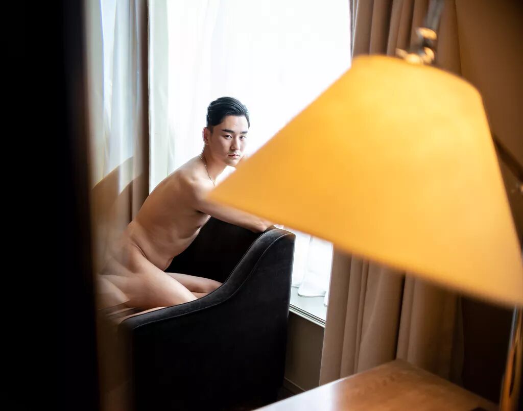 A nude gentleman kneels on a chair in front of a bright window