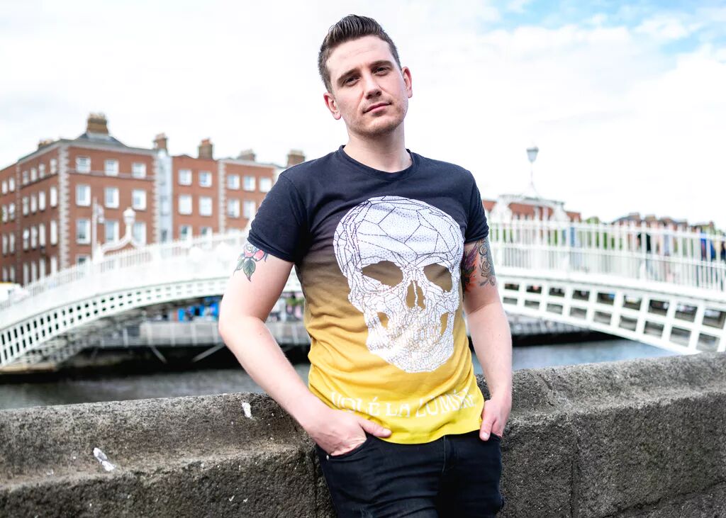 Young man in a colorful graphic tee with a skull on it leans casually against a stone railing in front of a river