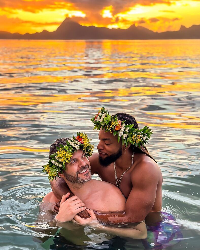 Emile and AJ wearing flower crowns embracing each other in the ocean at sunset.