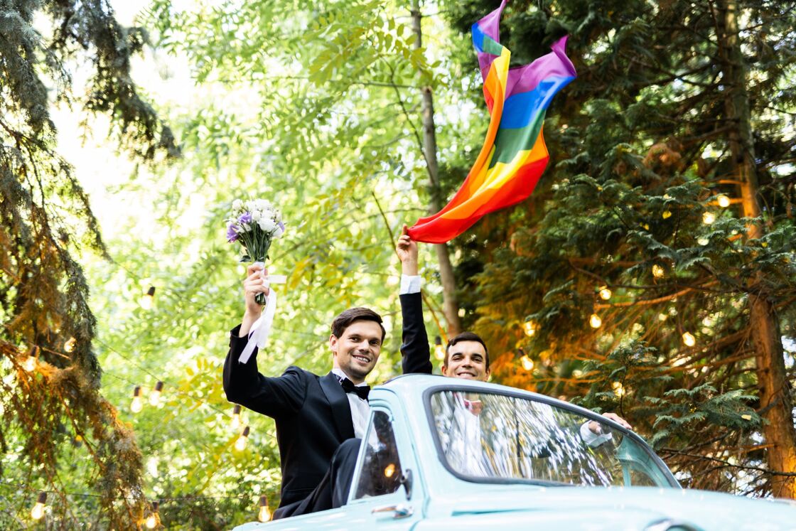 a cay couple holding a rainbow pride flag in the air while driving off to their honeymoon celebration