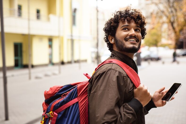 Smiling Indian tourist with backpack using smartphone on urban street