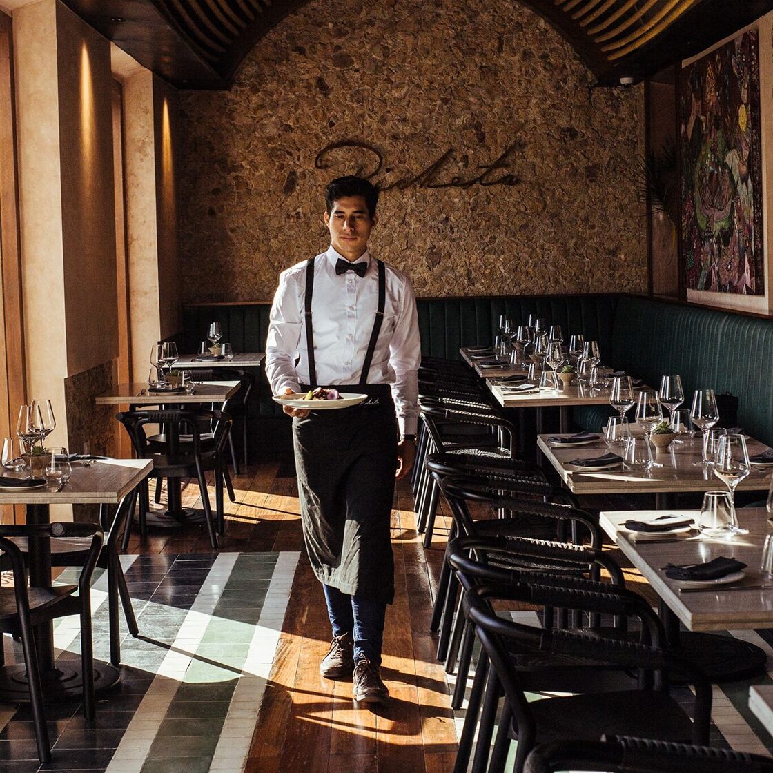 A handsome man in a white shirt, black boy tie, and suspenders approaches the camera with a plate of food.