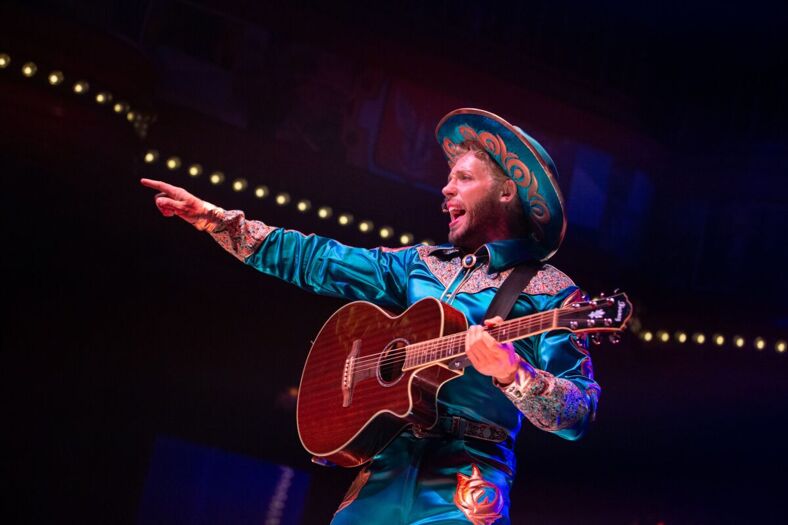A guitar player in a bright blue, shiny cowboy outfit smiles and points 