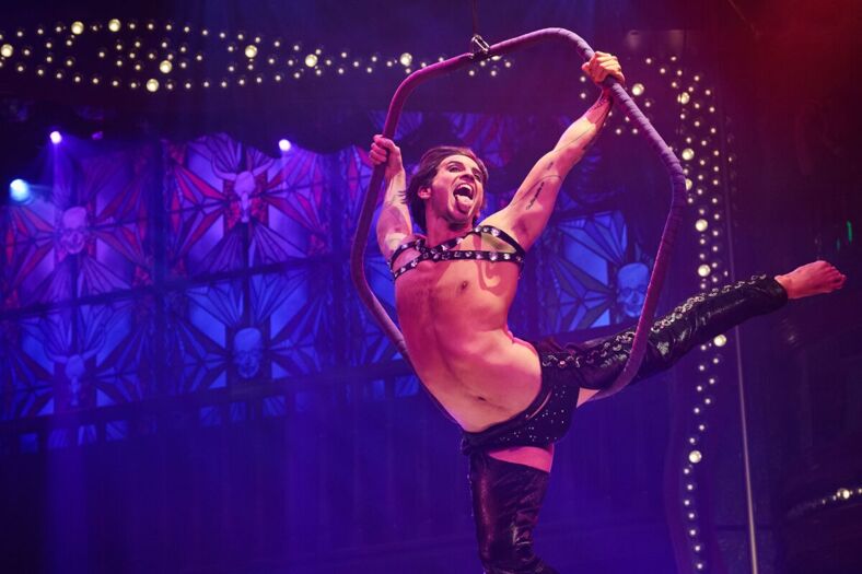 A handsome, shirtless aerial dancer dramatically poses while flying and sticking out his tongue