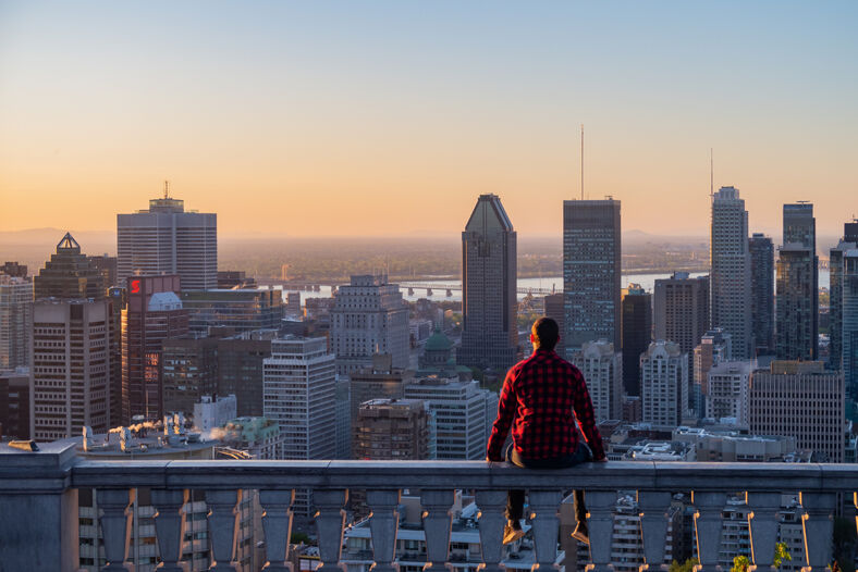 A man wearing a flannel shirt sits on a rail overlooking Montreal's skyline.