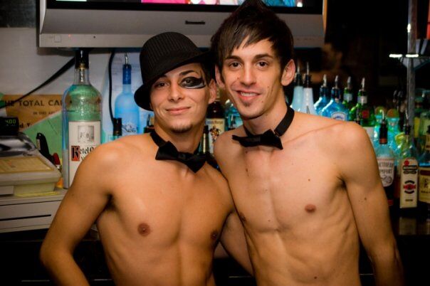 Two shirtless bartenders smile at the camera. Photo via: The Escape Lounge