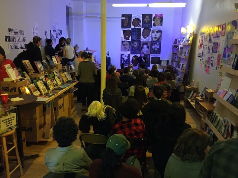An author reads an excerpt from their latest book at the front of a crowded room. 