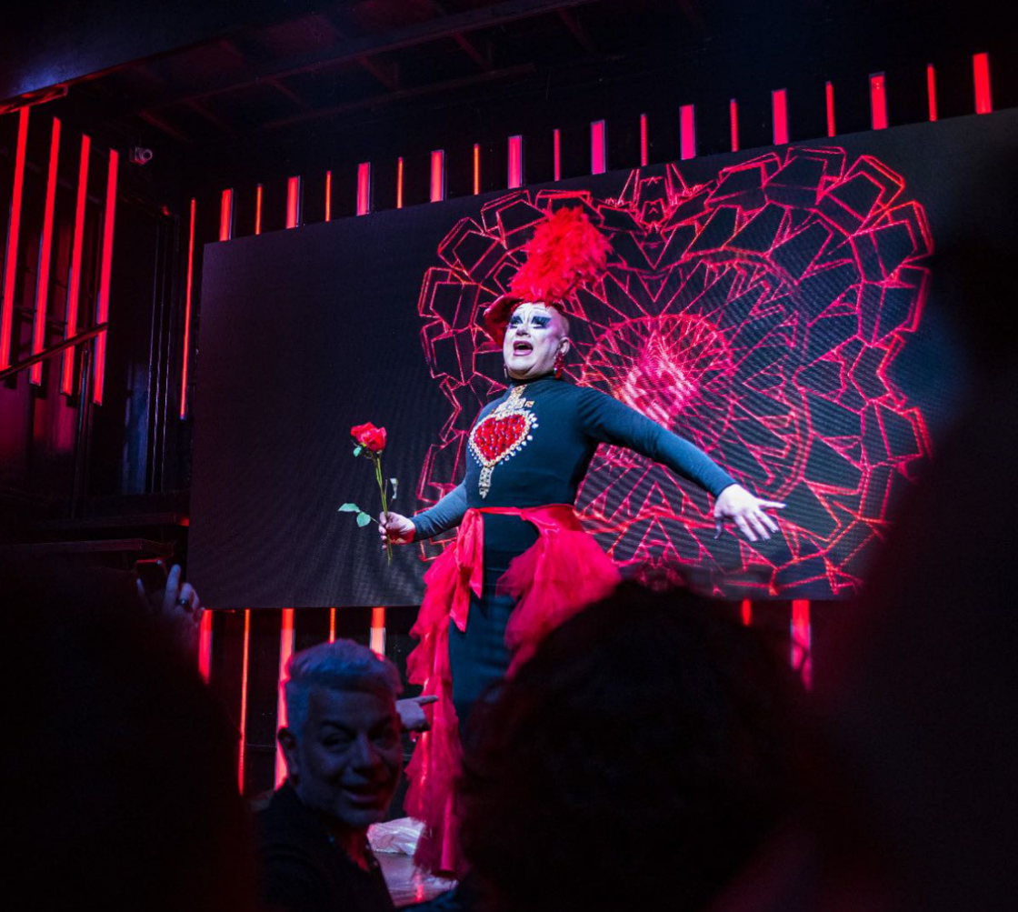 A drag queen wearing a black gown with a red hear on the chest and a red sash with a big red hat performs.