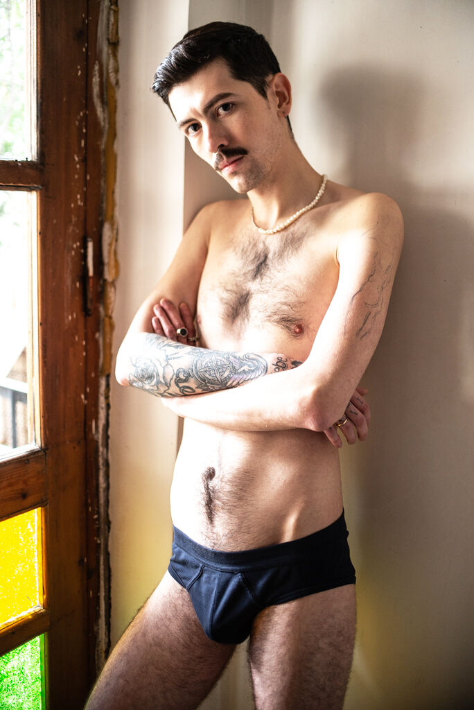 A shirtless tattooed man stands by a window wearing black underwear and pearl necklace.