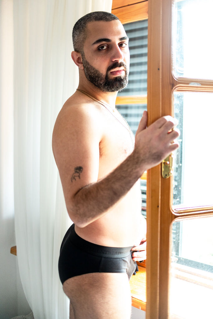 A shirtless man in black underwear stands by an open window.