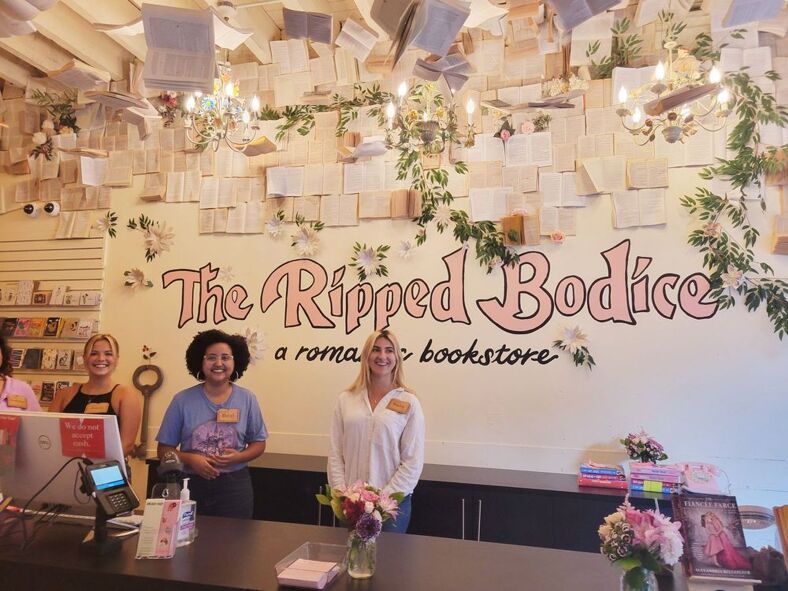 LGBTQ+ bookstores: A picture of three employees at the register of The Ripped Bodice in Brooklyn, New York. They all appear happy and smiling. Above them are pages from books hanging from the ceiling. 