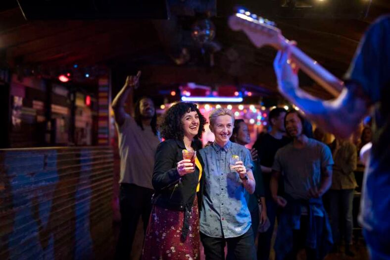 A lesbian couple embrace while holding cocktails. They are smiling at a musician on the stage. Photo via Visit Austin