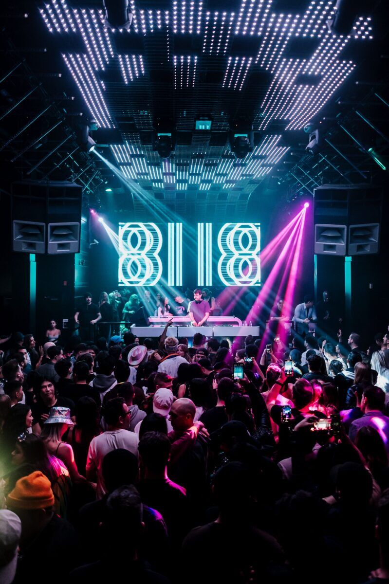 packed dance floor. At the center is a DJ booth with a huge neon sign behind it and purple lights. Photo via Celebrities Nightclub