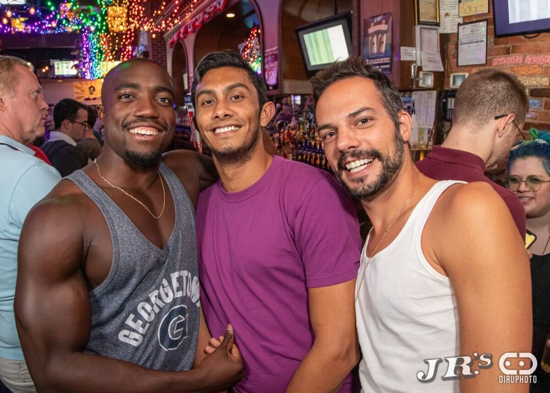 Three handsome men smile at the camera while posing in front of a crowded bar. Photo via JR's Bar